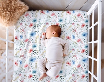 Op de afbeelding: Een baby slaapt op een wit ledikantlaken met een bloemenpatroon met roze, blauwe en groene aquarelbloemen.