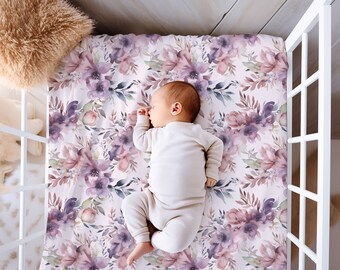 Op de afbeelding: Een baby slaapt op een wit ledikantlaken met een aquarel bloemenpatroon in tinten paars, roze en groen.