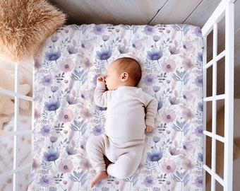 Op de afbeelding: Een baby slaapt op een wit ledikantlaken met een aquarel bloemenpatroon in tinten paars, roze en grijs.