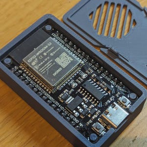Könnte beinhalten: Ein offenes, dunkelgraues rechteckiges elektronisches Gerät mit einer Leiterplatte und einem abgenommenen Deckel. Die Leiterplatte hat verschiedene Komponenten, darunter einen Chip mit der Bezeichnung ESP32-WROOM-32. Der Deckel hat eine Reihe paralleler Schlitze.