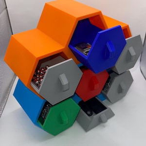 Pode incluir: Uma unidade de armazenamento modular composta por compartimentos hexagonais interconectados em laranja, azul, vermelho e verde. Várias gavetas hexagonais cinzentas estão parcialmente abertas, revelando pequenos itens como esferas de metal e componentes.