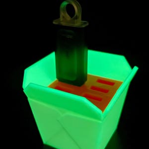 Op de afbeelding: Een lichtgevende groene container met een donkergroen rechthoekig object en een transparante ring. De container heeft een oranje binnenkant met sleuven. De achtergrond is zwart.