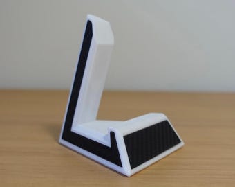 Op de afbeelding: Een witte telefoonstandaard met een zwart carbonfiber ontwerp op de basis. De standaard is naar links gekanteld en heeft een verhoogde lip aan de bovenkant om een telefoon vast te houden.