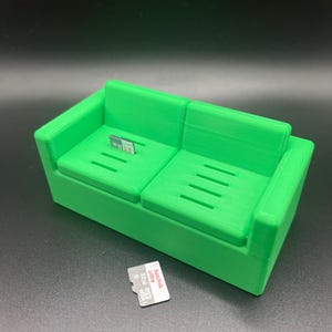 Può includere: Un organizzatore di archiviazione per schede di memoria a forma di divano stampato in 3D di colore verde. Il divano ha due posti a sedere ed è progettato per contenere più schede di memoria. Una piccola scheda di memoria è mostrata davanti al divano.