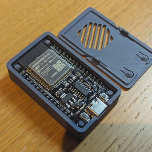 Könnte beinhalten: Ein dunkelgraues rechteckiges elektronisches Gerät mit offenem Deckel, das interne Komponenten freilegt. Das Gerät hat eine Leiterplatte mit verschiedenen Chips, Anschlüssen und dem Text "ESP32-WROOM-32". Der Deckel hat eine Reihe paralleler Schlitze.