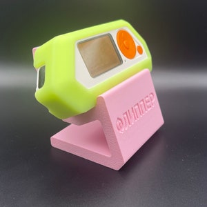 May include: A lime green and white electronic device with an orange button is displayed on a pink stand. The stand has the word "ОЛИМПЕЦ" printed on it.