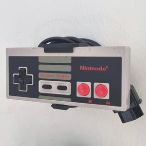 Op de afbeelding: Een grijze en zwarte Nintendo Entertainment System-controller met rode knoppen en een zwart snoer. De controller is gemonteerd aan een witte muur.