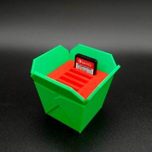 May include: A green and red 3D printed container shaped like a takeout box with a Nintendo Switch game cartridge inside.