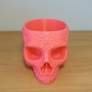 Könnte beinhalten: Ein rosafarbener Schädel-Blumentopf mit komplizierten Wirbelmustern. Der Topf ist aus einem glatten, plastikähnlichen Material gefertigt.
