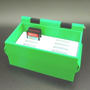 Könnte beinhalten: Eine hellgrüne, 3D-gedruckte Aufbewahrungsbox für Videospielkassetten. Die Box hat einen weißen Innenraum mit Schlitzen für Spielkassetten und einen Klappdeckel. Zwei Spielkassetten sind sichtbar. Für Nintendo Switch-Spielkassetten.