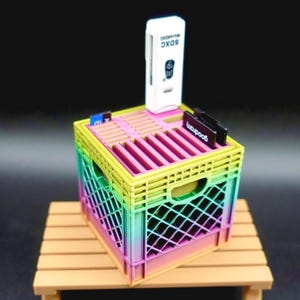 Könnte beinhalten: Eine Miniatur-Kiste in Regenbogenfarben mit einem weißen SDXC-Kartenleser und mehreren schwarzen und blauen Speicherkarten. Die Kiste steht auf einem kleinen Holztisch, mit dunklem Hintergrund.