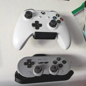 Puede incluir: Dos mandos de videojuegos montados en una pared. El mando superior es blanco con botones negros y un logotipo de Xbox negro. El mando inferior es gris con botones negros y el texto "8BitDo SN30 Pro" impreso en él.