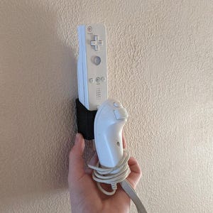 Puede incluir: Un mando de Wii blanco y un mando de Wii MotionPlus blanco están montados en una pared con una correa de velcro negra. Los mandos están conectados por un cable blanco.