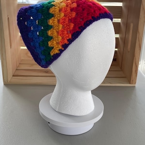 May include: A rainbow crochet headband with a purple, blue, green, yellow, orange, and red color pattern. The headband is displayed on a white mannequin head.