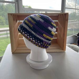 May include: A crocheted beanie hat with stripes of purple, black, white, and yellow.