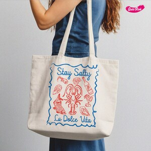 以下が含まれることがあります： 青い縁取りと「Stay Salty La Dolce Vita」という青い文字が書かれた白いキャンバス地のトートバッグ。バッグには、ロブスター、魚、その他のシーフードの赤いイラストが描かれている。