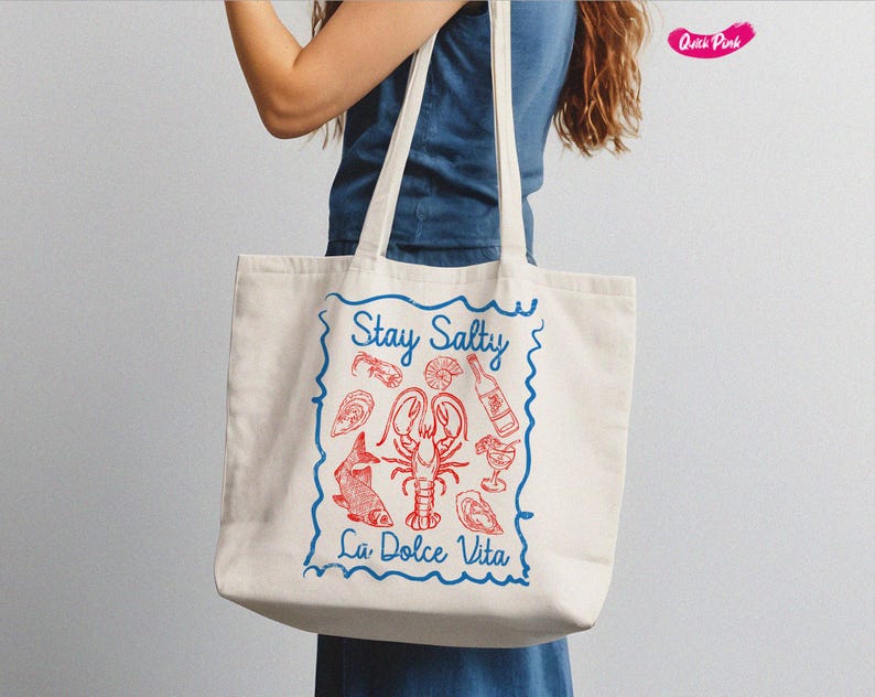 以下が含まれることがあります： 青い縁取りと「Stay Salty La Dolce Vita」という青い文字が書かれた白いキャンバス地のトートバッグ。バッグには、ロブスター、魚、その他のシーフードの赤いイラストが描かれている。