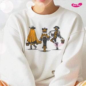 May include: White sweatshirt featuring a Halloween-themed graphic. The design depicts three figures: a ghost, a pumpkin-headed cowgirl, and a skeleton, each holding a trick-or-treat bucket. The figures are in a line, walking forward.