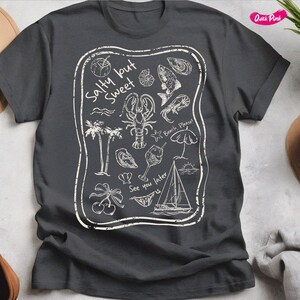 May include: A dark gray t-shirt with a white graphic design. The design features a variety of beach-themed illustrations, including a lobster, a sailboat, a cocktail glass, and the text "Salty but Sweet".