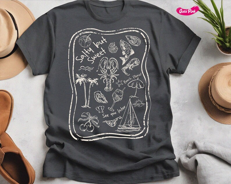 May include: A dark gray t-shirt with a white graphic design. The design features a variety of beach-themed illustrations, including a lobster, a sailboat, a cocktail glass, and the text "Salty but Sweet".