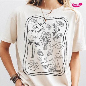 May include: White t-shirt with a black graphic print of a beach scene. The graphic includes a lobster, palm trees, a sailboat, a seashell, a fish, and the text "Salty but Sweet".