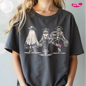 May include: Dark gray t-shirt featuring a Halloween-themed graphic. The design depicts three figures in cowboy attire, including a ghost, a pumpkin-headed person, and a skeleton, each holding a jack-o'-lantern bucket. The shirt has a crew neck and short sleeves.