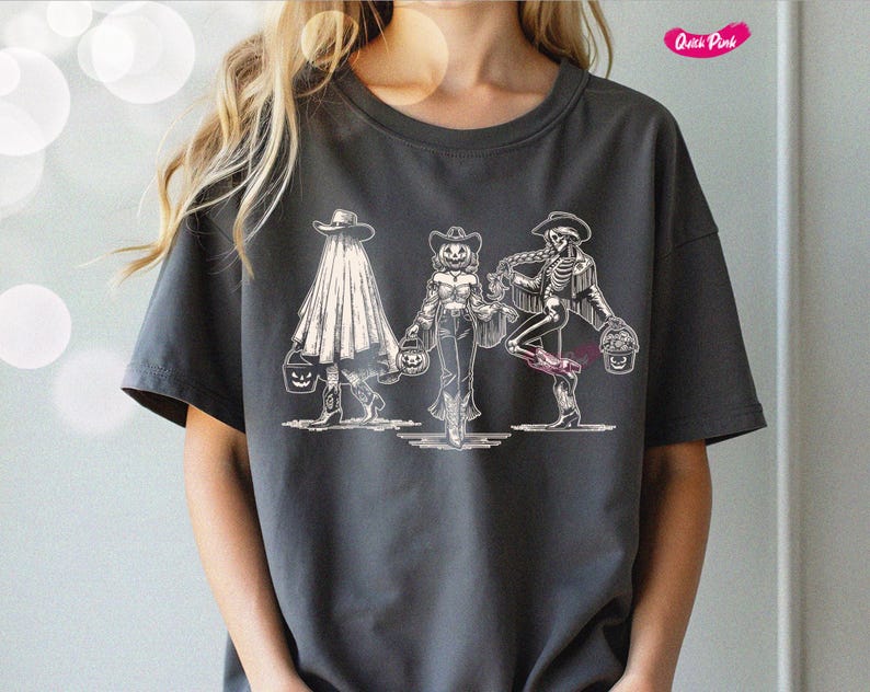 May include: Dark gray t-shirt featuring a Halloween-themed graphic. The design depicts three figures in cowboy attire, including a ghost, a pumpkin-headed person, and a skeleton, each holding a jack-o'-lantern bucket. The shirt has a crew neck and short sleeves.