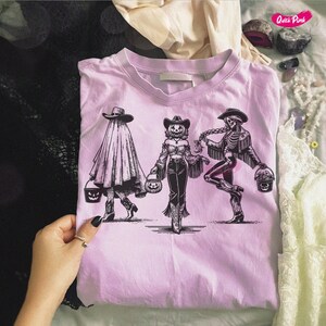 May include: A light pink t-shirt featuring a black and white illustration of three figures in Halloween costumes. The figures are holding pumpkin-shaped buckets. The figures are a ghost, a scarecrow, and a skeleton, all wearing cowboy hats.