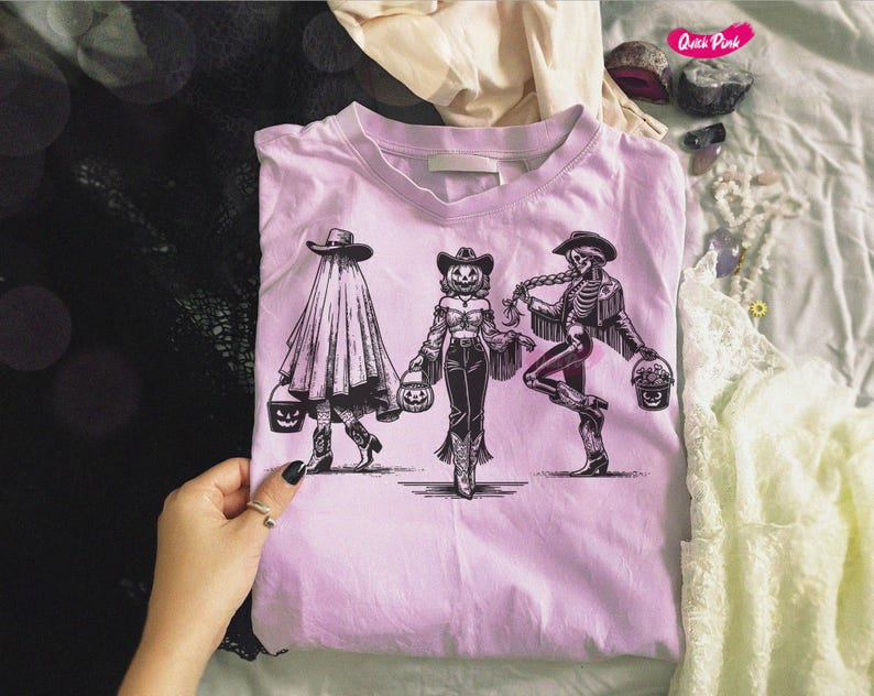 May include: A light pink t-shirt featuring a black and white illustration of three figures in Halloween costumes. The figures are holding pumpkin-shaped buckets. The figures are a ghost, a scarecrow, and a skeleton, all wearing cowboy hats.