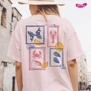 Pode incluir: T-shirt rosa claro com quatro desenhos de selos postais com ilustra&ccedil;&otilde;es de frutos do mar. Os selos incluem imagens de peixes, uma lagosta, um caranguejo com camar&otilde;es e ostras. As palavras "FIRST CLASS" s&atilde;o impressas no canto superior direito.