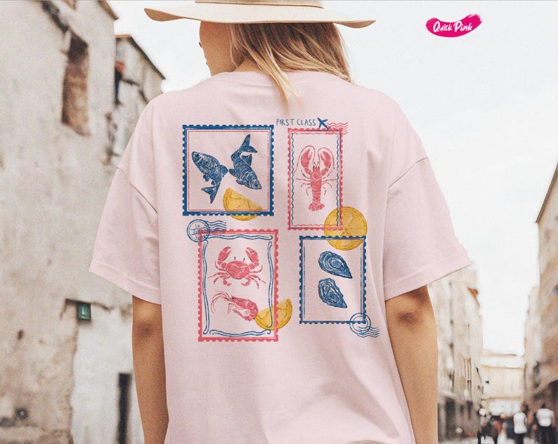 Pode incluir: T-shirt rosa claro com quatro desenhos de selos postais com ilustra&ccedil;&otilde;es de frutos do mar. Os selos incluem imagens de peixes, uma lagosta, um caranguejo com camar&otilde;es e ostras. As palavras "FIRST CLASS" s&atilde;o impressas no canto superior direito.