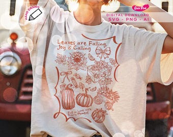 May include: A light beige t-shirt with a fall-themed graphic. The graphic includes the text "Leaves are Falling, Joy is Calling" and illustrations of pumpkins, leaves, and a sunflower. The shirt also has the text "Fall in Love with Every Moment."