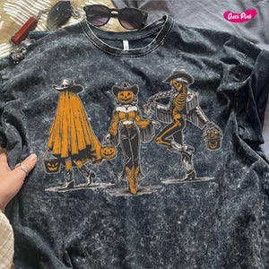 May include: Dark gray t-shirt with a Halloween-themed graphic. The graphic features three figures: a ghost, a pumpkin-headed cowgirl, and a skeleton cowboy, each holding a trick-or-treat bucket. The shirt has a textured, washed appearance.
