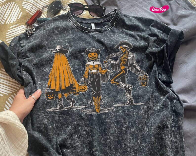 May include: Dark gray t-shirt with a Halloween-themed graphic. The graphic features three figures: a ghost, a pumpkin-headed cowgirl, and a skeleton cowboy, each holding a trick-or-treat bucket. The shirt has a textured, washed appearance.