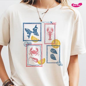 Pode incluir: Camiseta creme com quatro desenhos de selos postais com ilustra&ccedil;&otilde;es de frutos do mar. Os selos retratam peixes, uma lagosta, um caranguejo com camar&otilde;es e ostras. Cada selo &eacute; emoldurado em azul ou rosa com o texto First Class.