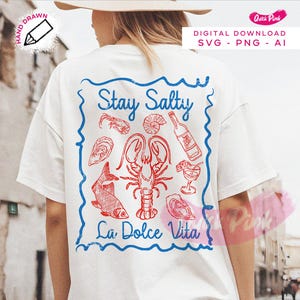 以下が含まれることがあります： 「Stay Salty La Dolce Vita」と書かれた青と赤のグラフィックデザインが施された白いTシャツ。グラフィックには、ロブスター、魚、ワインボトル、その他のシーフードのイメージが含まれています。