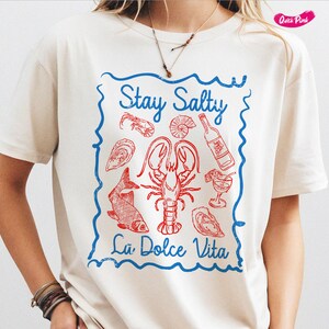 以下が含まれることがあります： 「Stay Salty La Dolce Vita」と書いてある、青と赤のグラフィックプリントが施された白いTシャツ。グラフィックには、ロブスター、魚、貝殻、ワインボトルが描かれています。