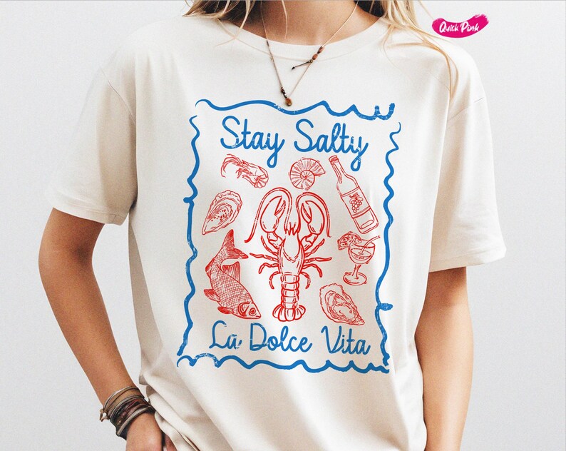 以下が含まれることがあります： 「Stay Salty La Dolce Vita」と書いてある、青と赤のグラフィックプリントが施された白いTシャツ。グラフィックには、ロブスター、魚、貝殻、ワインボトルが描かれています。
