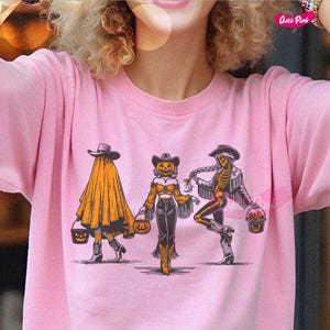 May include: Pink t-shirt featuring a Halloween-themed graphic. The design includes three figures: a ghost, a scarecrow, and a skeleton, each holding a pumpkin bucket. The figures are wearing cowboy hats and boots.