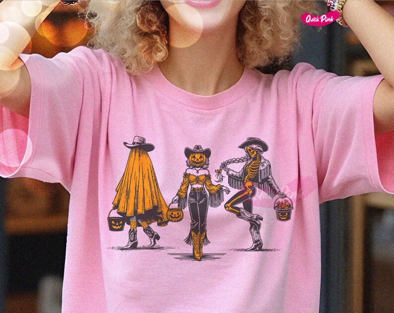 May include: Pink t-shirt featuring a Halloween-themed graphic. The design includes three figures: a ghost, a scarecrow, and a skeleton, each holding a pumpkin bucket. The figures are wearing cowboy hats and boots.