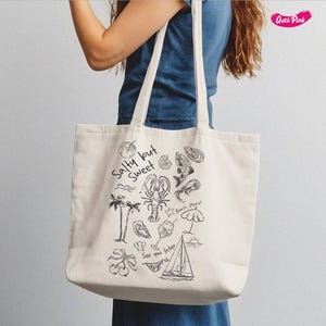 May include: A white canvas tote bag with black illustrations of a lobster, fish, palm trees, and other beach-themed items. The text "Salty but Sweet" is printed on the bag.