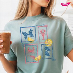 Pode incluir: Uma camiseta azul clara com quatro desenhos de selos postais representando frutos do mar. Os selos mostram peixes, uma lagosta, um caranguejo e ostras, com fatias de lim&atilde;o. O texto "FIRST CLASS" &eacute; impresso acima dos selos.
