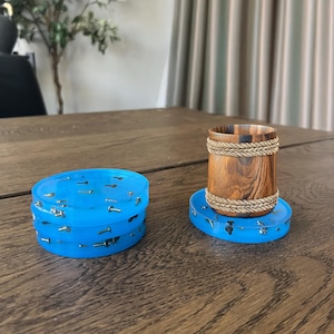 May include: A stack of three translucent blue circular resin coasters embedded with small metal screws, and a single matching coaster supporting a cylindrical wooden cup featuring two decorative braided rope bands around its exterior.