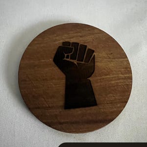 May include: A round wooden coaster with a dark brown finish. The coaster features a black fist graphic in the center. The text "SOLIDARITY WITH EVERY SIP" is at the bottom in a black rectangle.