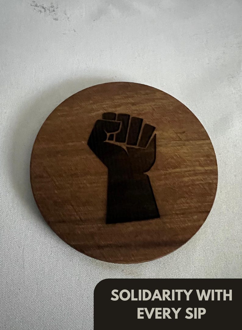 May include: A round wooden coaster with a dark brown finish. The coaster features a black fist graphic in the center. The text "SOLIDARITY WITH EVERY SIP" is at the bottom in a black rectangle.