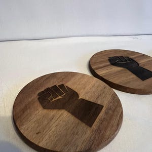 May include: Two round wooden coasters with a fist design engraved on them. The coasters are made of a light brown wood and have a dark brown fist design. The text "ENGRAVED WITH POWER" is at the bottom.