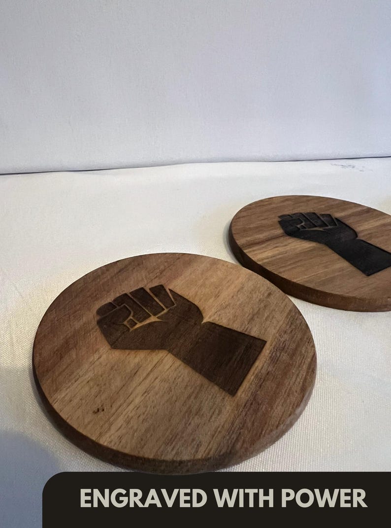 May include: Two round wooden coasters with a fist design engraved on them. The coasters are made of a light brown wood and have a dark brown fist design. The text "ENGRAVED WITH POWER" is at the bottom.