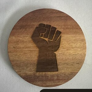 May include: Round wooden coaster with a carved fist design. The coaster is made of light brown wood and features the text "HANDMADE & UNAPOLOGETIC" in a black rectangle at the bottom. The fist is a symbol of solidarity and resistance.