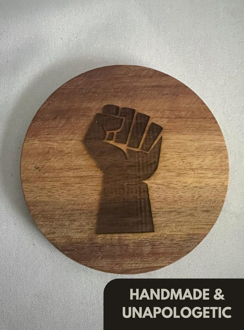 May include: Round wooden coaster with a carved fist design. The coaster is made of light brown wood and features the text "HANDMADE & UNAPOLOGETIC" in a black rectangle at the bottom. The fist is a symbol of solidarity and resistance.