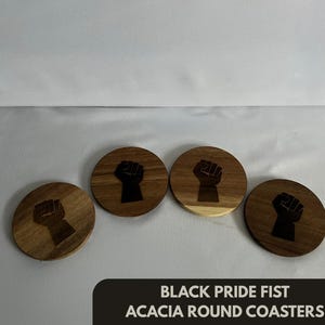 May include: Four round acacia wood coasters with a black fist design. The coasters are arranged on a light surface. The text "BLACK PRIDE FIST ACACIA ROUND COASTERS" is at the bottom.